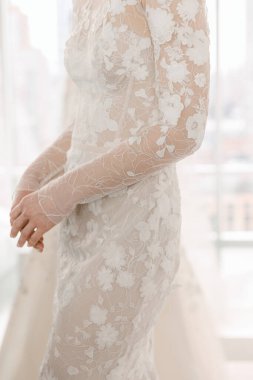 Gelin, güneş ışığıyla dolu bir odada dikilirken çiçek desenleriyle süslenmiş muhteşem dantel elbisesini sergiliyor..