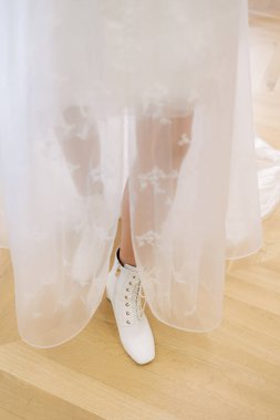 Bir gelin göz kamaştırıcı dantelli gelinliğini ve şık beyaz botlarını parlak bir odada sergiliyor..