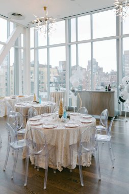 Parlak bir olay alanı beyaz masa örtüleri ve şeffaf sandalyeleri olan yuvarlak masaları içerir ve şehrin ufuk çizgisi görünümünü sağlar.