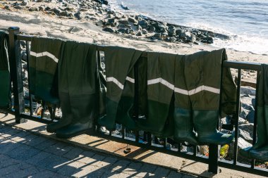 Islak balıkçı kıyafeti bir çite asılır, sahilde güneşte kurur, dalgalar yavaşça kıyıya vurur..