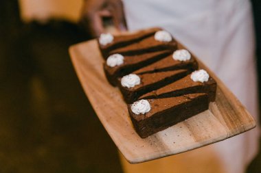 Bir insan, üzerinde krem şantili beş çikolatalı browni olan ahşap bir tabağı sunar..