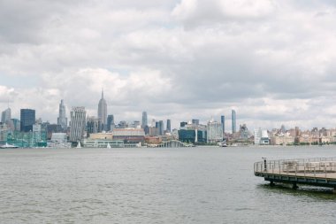 Bulutlu bir gökyüzü ve uzak bir rıhtımla sudan görünen hareketli bir şehrin silüeti..