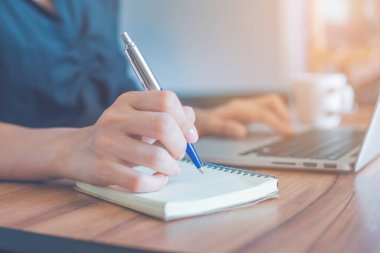 Kadın kalemle deftere yazı yazıyor ve ofiste çalışmak için dizüstü bilgisayar kullanıyor. Web pankartı.