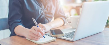 Kadın kalemle deftere yazı yazıyor ve ofiste çalışmak için dizüstü bilgisayar kullanıyor. Web pankartı.