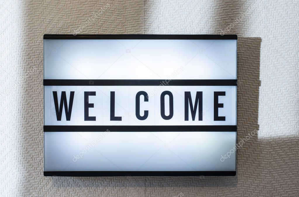 Message Welcome on illuminated board. Welcoming concept with text. Daylight from window. Room interior. Black letters welcome on white wallpaper wall.