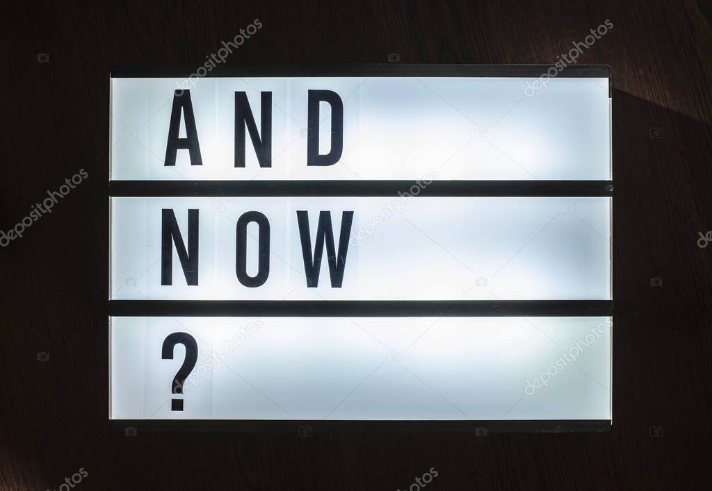 Message And Now? on illuminated board. Question about decision concept with text. Daylight from window. Room interior. Black letters And Now on dark wallpaper wall.