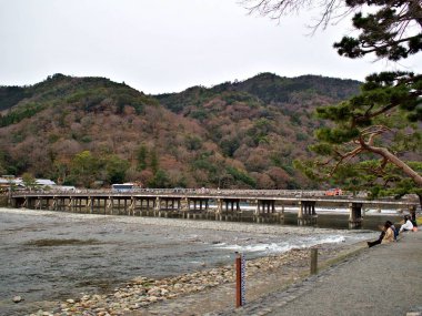 İnsanlar nehir bankında rahatlatıcı oturan ve Arashiyama erken Bahar sırasında güzel bir tarihi Togetsu-kyo köprü görme, Kyoto, Japonya