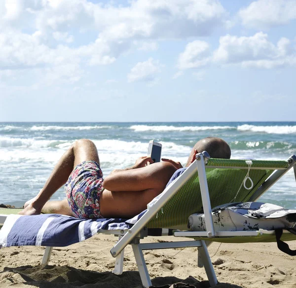 Людина Пляжі Лежачи Солярію Дивлячись Смартфон Тлі Двох Чоловіків Споглядати Ліцензійні Стокові Зображення