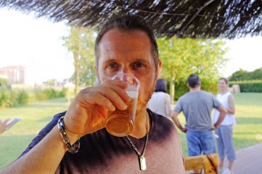 Açık bir barda bira içen adam. Arka planda bazı arkadaşlar zevk. Plastik camda bira.