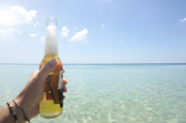 Erkek el güneşli bir gökyüzü ve kristal berraklığında denize karşı bir şişe bira tutan. Tatil konsepti.