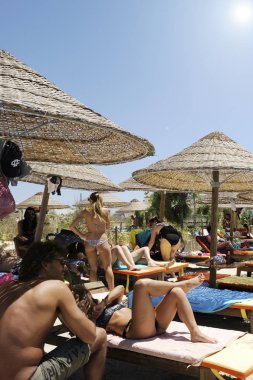 Mavi bir gökyüzü ve beyaz bulutlar üzerinde beyaz saman şemsiyeile güzel plaj tesisi. Arka planda bazı insanlar sahilde eğleniyor. Plaj hayatı ve yaşam tarzı kavramı.