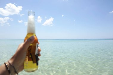 Erkek el güneşli bir gökyüzü ve kristal berraklığında denize karşı bir şişe bira tutan. Tatil konsepti.