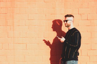 Günlük giysili ve cep telefonlu güneş gözlüklü bir adam açık havada kırmızı duvarda dikilmekten zevk alan genç beyaz bir adam, teknoloji cihazlarıyla boş zaman aktivitesi, kırmızı duvar arka planı ve uzay kopyası.