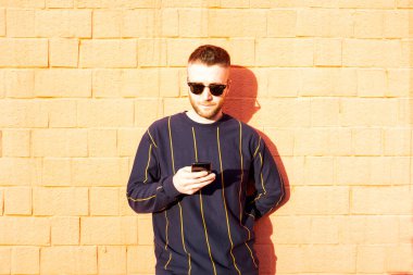 Cep telefonlu sıradan giyimli bir adam - açık havada kırmızı duvarda duran beyaz bir adam ve akıllı telefonlu videolar ya da sosyal ağlar görüyor - teknoloji cihazlarıyla boş zaman aktivitesi - ışık filtresi