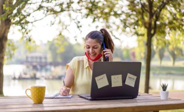 Young woman wearing open face mask calling with smartphone while working with laptop outdoors - Smiling female freelancer talking on cell phone and sitting with laptop in park during coronavirus time clipart
