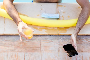 Havuz başında eğlenen genç bir adamın, akıllı telefonuyla arkadaşlarını araması Coronavirus salgını sırasında sağlıklı meyve suyu içmesi sosyal ilişki üzerinde Covid-19 etkisi, kollara odaklanması.