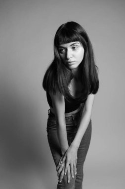 Siyah bluzlu güzel esmer bir kadının stüdyo portresi, yarı eğilmiş, iki koluyla dizine yaslanmış, düz gri bir arka plana bakıyor.