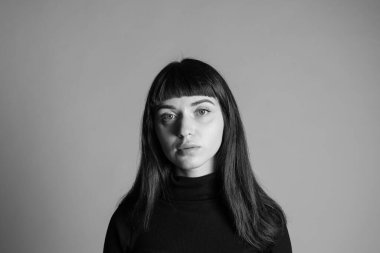 Güzel esmer bir kadının stüdyo portresini kapat, katlanmış siyah polo yaka kazak giyiyor, ciddi ciddi kameraya bakıyor, düz gri bir arka plana bakıyor.