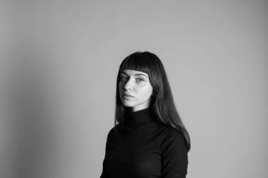 Güzel esmer bir kadının stüdyo portresi, katlanmış siyah polo yakalı bir süveter giyiyor, kameraya bakıyor, düz gri bir arka planda.