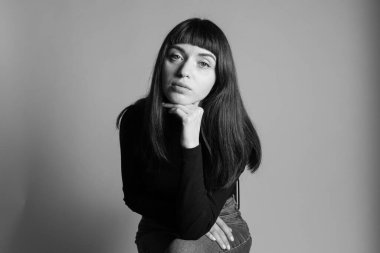 Güzel esmer bir kadının stüdyo portresi, katlanmış siyah polo yakalı bir süveter giyiyor, kameraya bakıyor, düz gri bir arka planda.