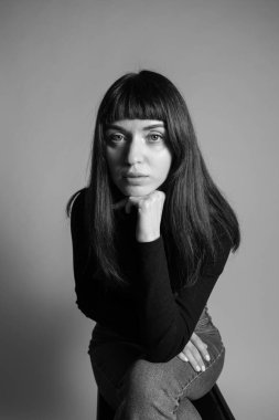 Güzel esmer bir kadının stüdyo portresi, katlanmış siyah polo yakalı bir süveter giyiyor, kameraya bakıyor, düz gri bir arka planda.