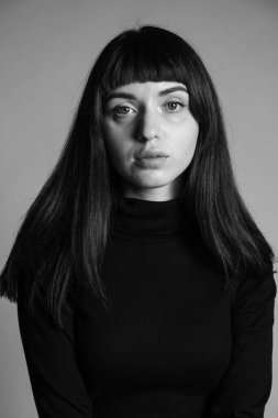 Güzel esmer bir kadının stüdyo portresi, katlanmış siyah polo yakalı bir süveter giyiyor, kameraya bakıyor, düz gri bir arka planda.