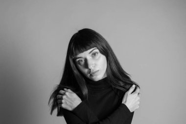 Güzel esmer bir kadının stüdyo portresini kapat, katlanmış siyah polo yaka kazak giyiyor, elleri omuzlarında, kameraya bakıyor, düz gri bir arkaplana bakıyor.