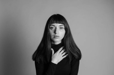 Güzel esmer bir kadının stüdyo portresini kapat, katlanmış siyah polo yakalı bir kazak giyiyor, elleri göğsünde, kameraya bakıyor, düz gri bir arka plana bakıyor.