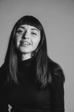 Güzel esmer bir kadının stüdyo portresini kapat, katlanmış siyah polo yaka kazak giyiyor, gülümsüyor, kameraya bakıyor, düz gri bir arka plana bakıyor.