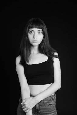 Siyah spagetti askılı güzel esmer bir kadının stüdyo portresi. Kollarını kavuşturmuş, kameraya bakıyor, düz siyah bir arkaplanda duruyor.