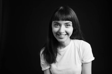 Beyaz tişörtlü esmer bir kadının stüdyo portresi. Siyah bir arka plana bakıp gülüyor.