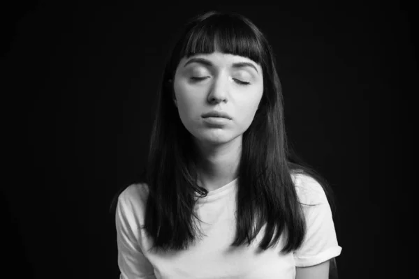 Studio portrait of a pretty brunette woman in a white blank t-shirt, sitting with closed eyes against a plain black background, looking at the camera