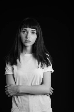 Beyaz, boş tişörtlü, kollarını kavuşturmuş, siyah bir arka planda kameraya bakan esmer bir kadının stüdyo portresi.