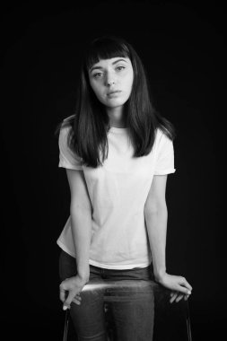 Beyaz tişörtlü güzel esmer bir kadının stüdyo portresi, bir sandalyenin arkasında, düz siyah bir arka planda, kameraya bakıyor.