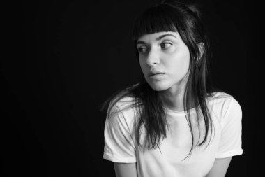 Beyaz, boş tişörtlü, siyah arka planda kameraya bakan esmer bir kadının stüdyo portresi.