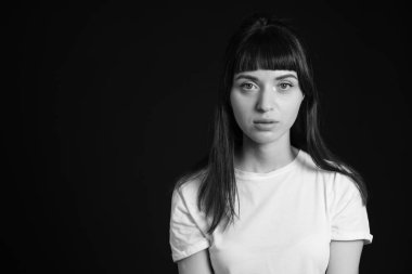 Beyaz, boş tişörtlü, siyah arka planda kameraya bakan esmer bir kadının stüdyo portresi.