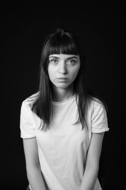 Beyaz, boş tişörtlü, siyah arka planda kameraya bakan esmer bir kadının stüdyo portresi.