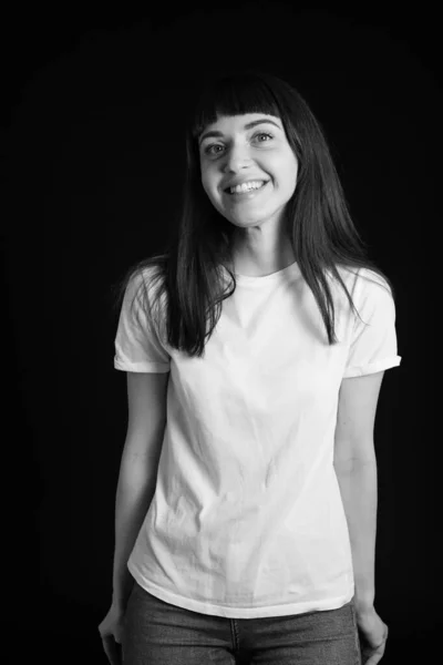 一个穿着白色空白T恤的漂亮的黑发女人站在工作室的画像 面对着一个普通的黑色背景微笑着 看着摄像机 — 图库照片