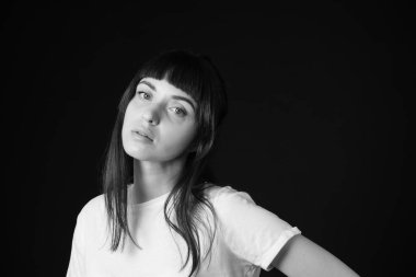 Beyaz, boş tişörtlü, siyah arka planda kameraya bakan esmer bir kadının stüdyo portresi.