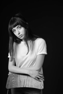 Beyaz, boş tişörtlü güzel esmer bir kadının stüdyo portresi. Sandalyenin arkasında duruyor. Kollarını çapraz yapıp, siyah arka planda kameraya bakıyor.