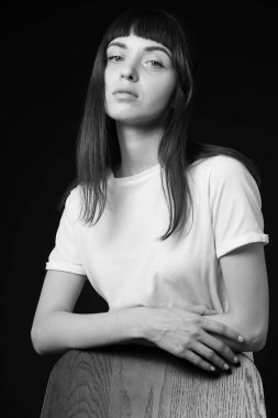 Beyaz, boş tişörtlü güzel esmer bir kadının stüdyo portresi. Sandalyenin arkasında duruyor. Kollarını çapraz yapıp, siyah arka planda kameraya bakıyor.