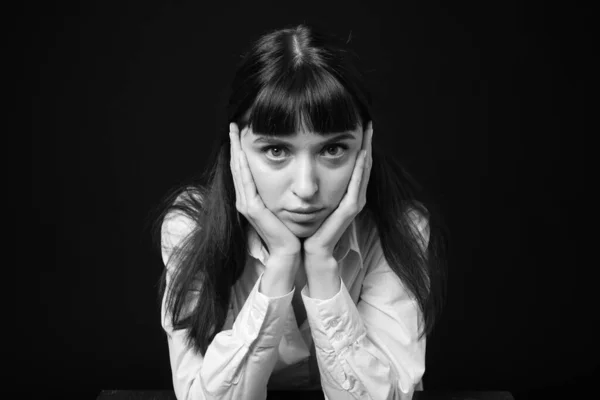 Studio portrait of a pretty brunette woman in a white shirt, sitting at the table, against a plain black background, seriously looking at the camera