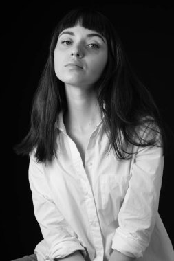 Beyaz gömlekli güzel esmer bir kadının stüdyo portresi, düz siyah arka planda oturuyor, kameraya bakıyor.