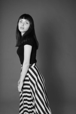 Siyah tişörtlü ve çizgili etekli güzel esmer bir kadının stüdyo portresi. Gri bir arka plana doğru yarım yamalak dönmüş, kameraya bakıyor.