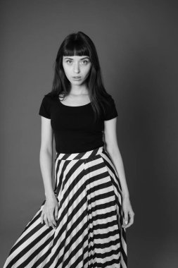 Siyah tişörtlü ve çizgili etekli güzel esmer bir kadının stüdyo portresi, düz gri bir arka planda kameraya bakıyor.