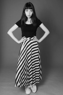 Siyah tişörtlü ve çizgili etekli güzel esmer bir kadının stüdyo portresi, düz gri bir arka planda kameraya bakıyor.