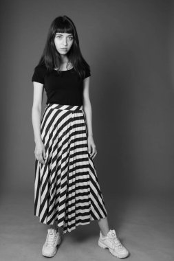 Siyah tişörtlü ve çizgili etekli güzel esmer bir kadının stüdyo portresi, düz gri bir arka planda kameraya bakıyor.