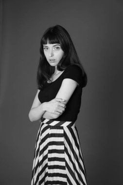 Siyah tişörtlü güzel esmer bir kadının stüdyo portresi, yarım yamalak duruyor, düz gri bir arka planda, kameraya bakıyor.