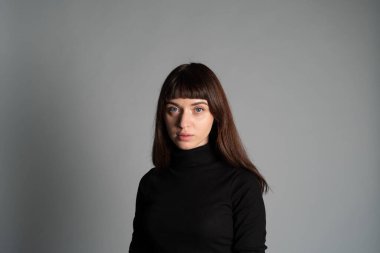 Güzel esmer bir kadının stüdyo portresi, katlanmış siyah polo yakalı bir süveter giyiyor, kameraya bakıyor, düz gri bir arka planda.