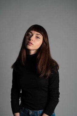 Güzel esmer bir kadının stüdyo portresi, katlanmış siyah polo yakalı bir süveter giyiyor, kameraya bakıyor, düz gri bir arka planda.